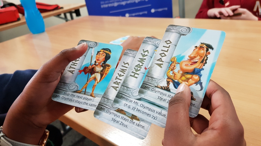 Close up of gaming cards from Zeus on the Loose in the hands of a child. Cards feature information and illustrations of Athena, Apollo, Hermes and Artemis.